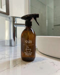 Shift reusable amber glass bathroom cleaner spray bottle with a black trigger on a white marble countertop.