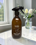 Shift amber glass multi-purpose spray bottle with black trigger on a white marble countertop near a flower vase.
