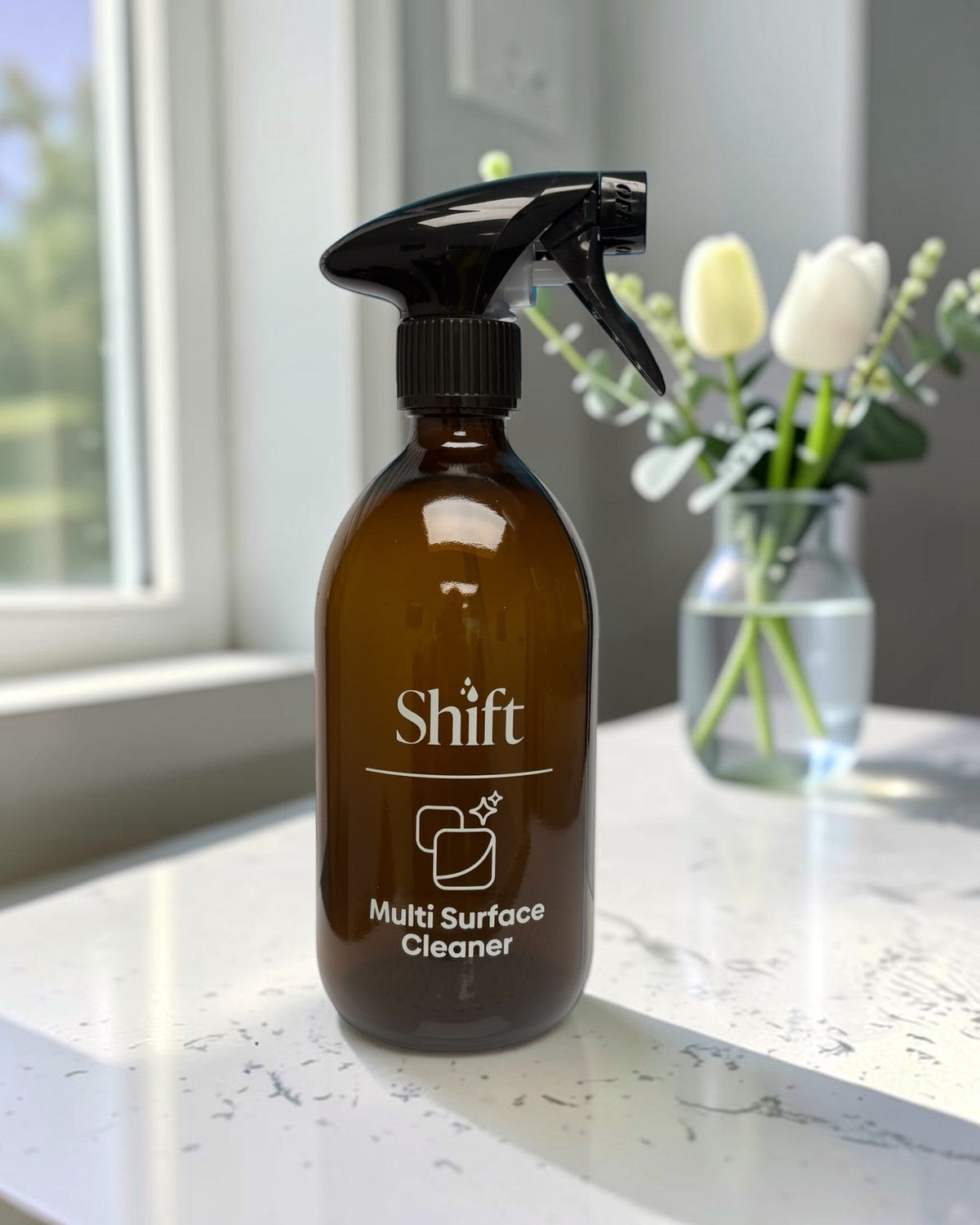 Shift amber glass multi-purpose spray bottle with black trigger on a white marble countertop near a flower vase.