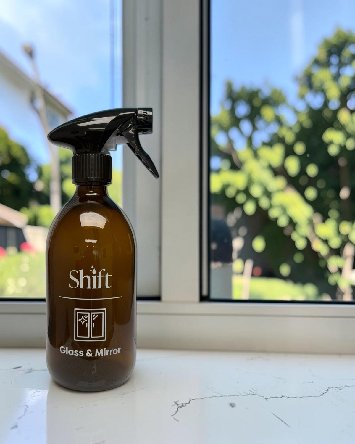 Shift amber glass spray bottle for glass and mirror cleaning with a black trigger nozzle on a white windowsill.